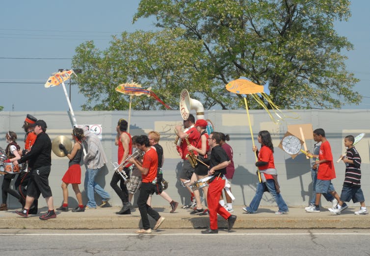 People march along a sidewalk playing instruments and holding signs featuring fish.
