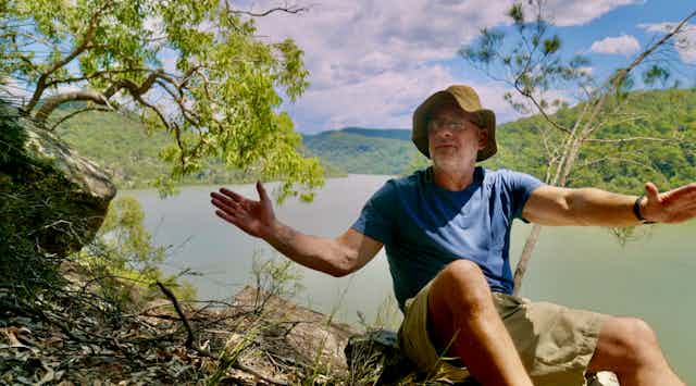 Tim Flannery in shorts and a t-shirt with outstretched arms, sitting above the Hawkesbury River in a still image from the documentary Climate Changers