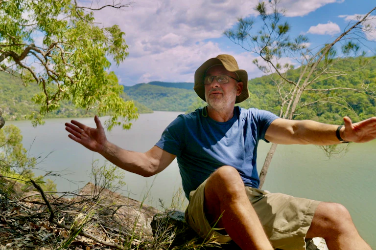Tim Flannery in shorts and a t-shirt with outstretched arms, sitting above the Hawkesbury River in a still image from the documentary Climate Changers