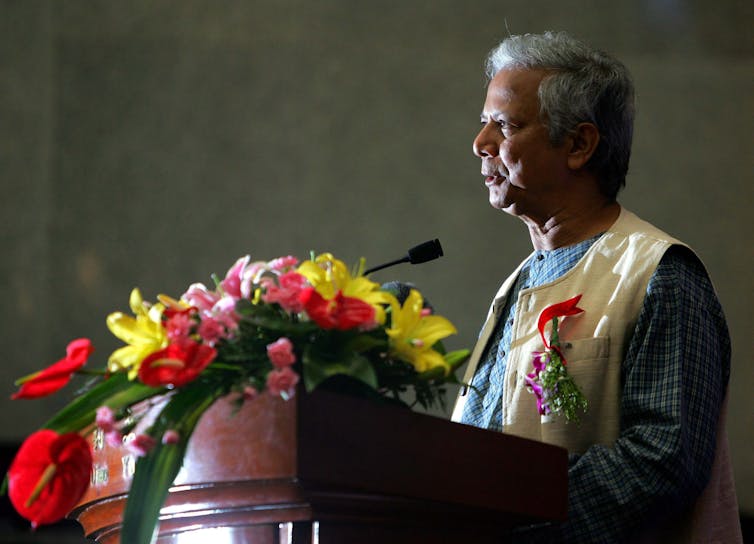 A grey-haired man speaks into a microphone behind a podium festooned with flowers.