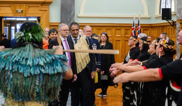 Prime Minister Albanese is walking into New Zealand Parliament alongside senior cultural advisor to Parliment Kura Moeahu. They are standing near Māori people doing a welcome ceremony.