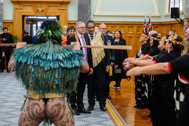 Prime Minister Albanese is walking into New Zealand Parliament alongside senior cultural advisor to Parliment Kura Moeahu. They are standing near Māori people doing a welcome ceremony.
