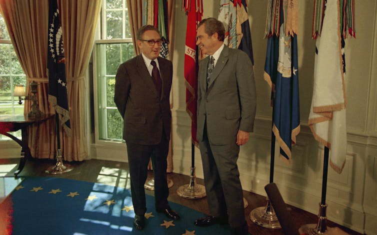 Two men in suits smile as they stand in an ornate office with flags behind them.