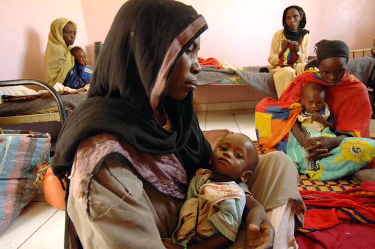 A Sudanese mother with scarf on her head is holding her malnourished son in a hospital.