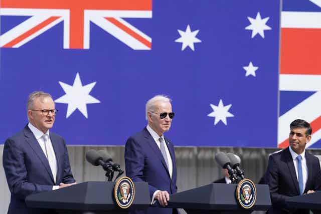 Three men, one in sunglasses, stand behind separate podiums in front of large flags.