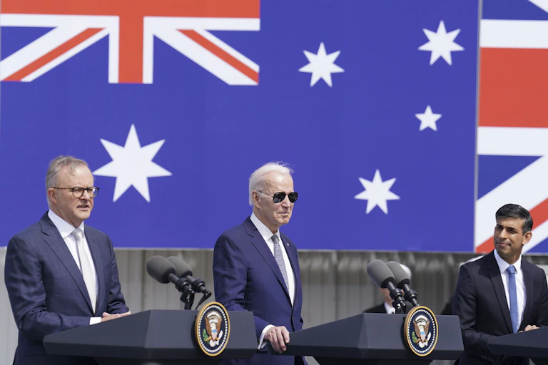 Three men, one in sunglasses, stand behind separate podiums in front of large flags.