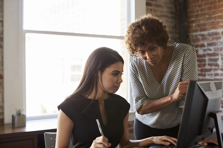 Woman stands over younger woman's desk as she works on computer