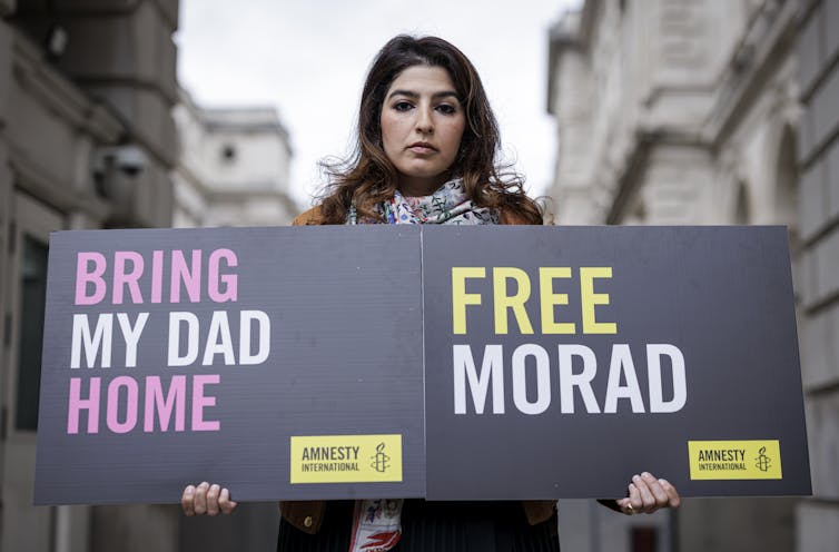 A woman standing outdoors holds two signs in her hands. One reads, 