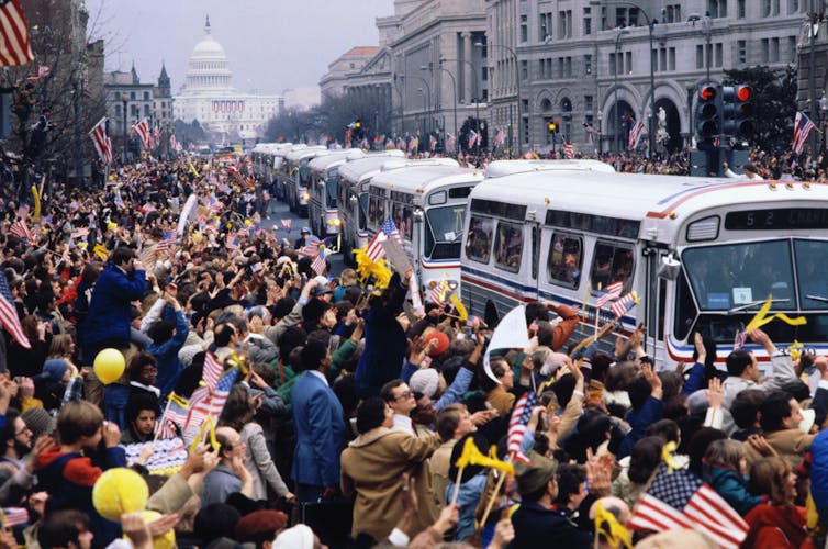 Hundreds of people stand on a sidewalk waving American flags and yellow ribbons at a line of passing buses.