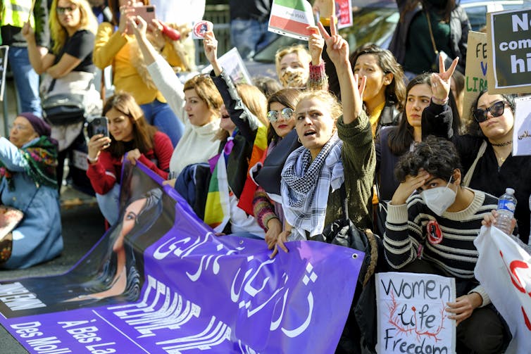 People, some sitting, others standing protest outside. Some hold placards while others use their hands to demonstrate the peace sign.