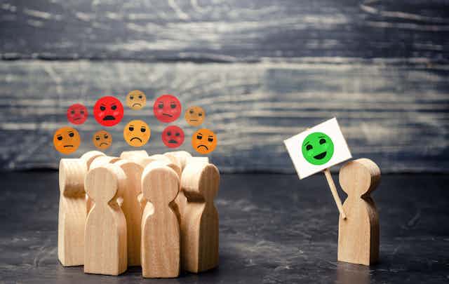 A graphic showing a group of unhappy people grouped together looking over to someone on their own showing a placard with a smiley face.