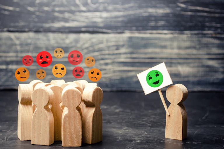 A graphic showing a group of unhappy people grouped together looking over to someone on their own showing a placard with a smiley face.