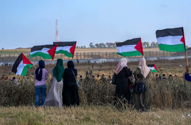 A row of women in headscarves wave Palestinian flags in a dusty field overlooking behind a barbed wire fence. 