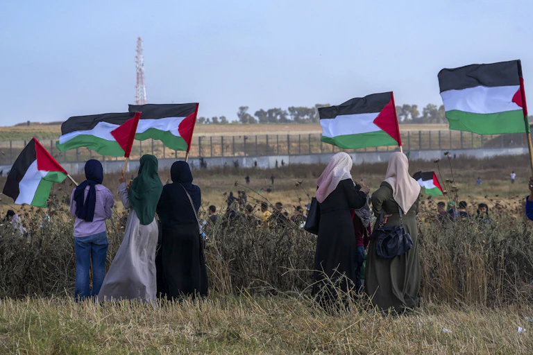 A row of women in headscarves wave Palestinian flags in a dusty field overlooking behind a barbed wire fence.