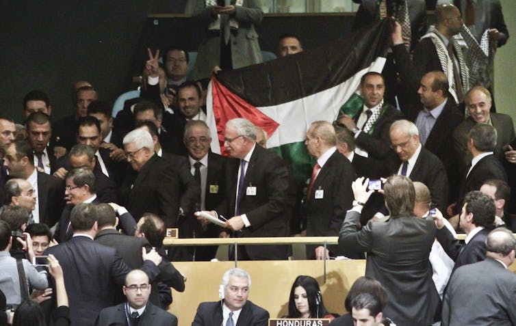 Men in suit in a conference hall with a large Palestinian flag being held behind them.