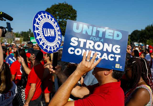 A sea of people wearing red shirts are shown rallying in an open space in Detroit. One prominently displays a blue sign that reads "UNITED FOR A STRONG CONTRACT"; behind him, another carries a UAW sign.