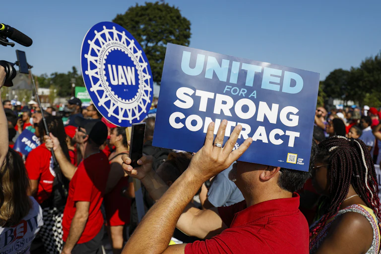 A sea of people wearing red shirts are shown rallying in an open space in Detroit. One prominently displays a blue sign that reads "UNITED FOR A STRONG CONTRACT"; behind him, another carries a UAW sign.