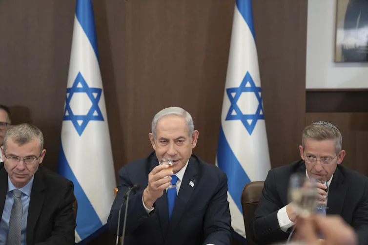 A man sitting at a table with two other men raises a glass with blue and white flags behind him.