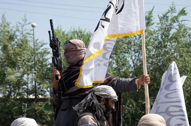 A man with his face covered holds a rifle in one hand a white flag in the other.