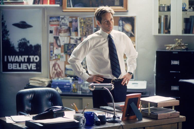 David Duchovny in an office wearing a black tie with a UFO poster behind him.