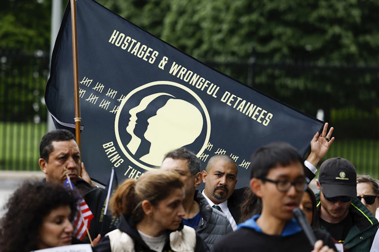 A crowd of people standing outside listen to someone speak as one man holds up a banner that reads, “Hostages & Wrongful Detainees.”