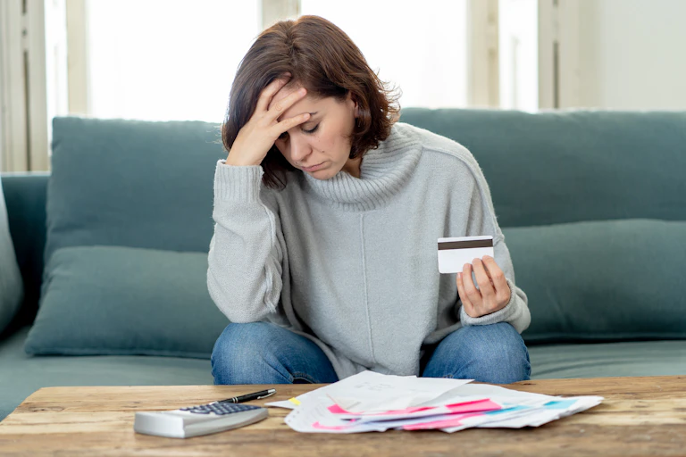 Woman with hand on head looking down at a pile of papers and a calculator, holding a credit card.