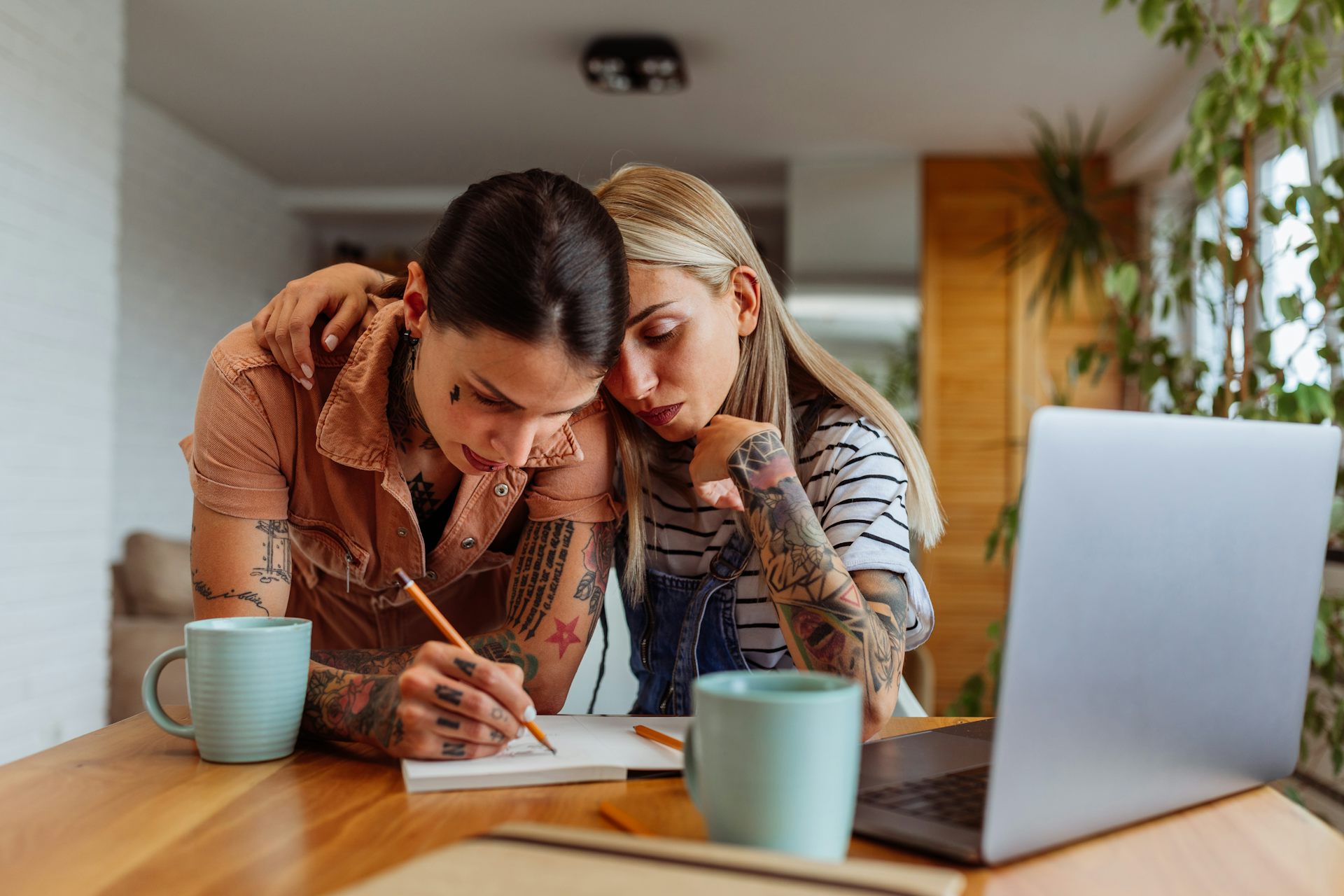 Two women with a pad, paper, laptop and mugs at kitchen table.