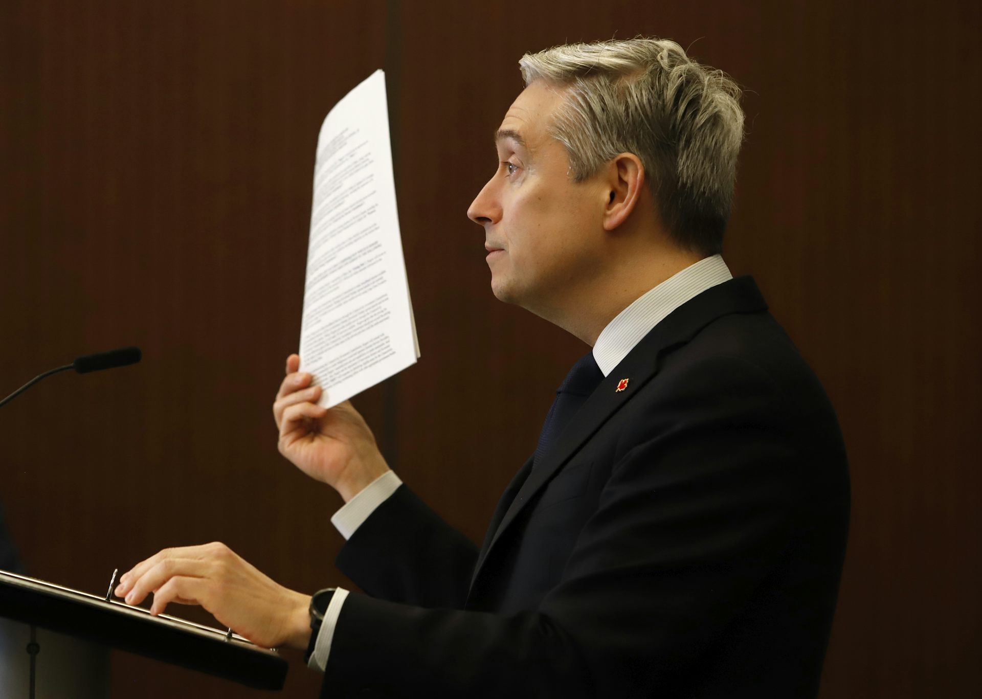 A man in profile holds up a bundle of papers from behind a podium