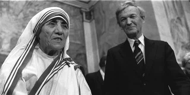 An elderly woman in a nun's habit stands nest to a grey-haired man.