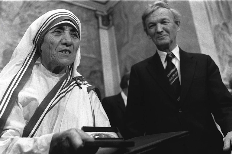 An elderly woman in a nun's habit stands nest to a grey-haired man.