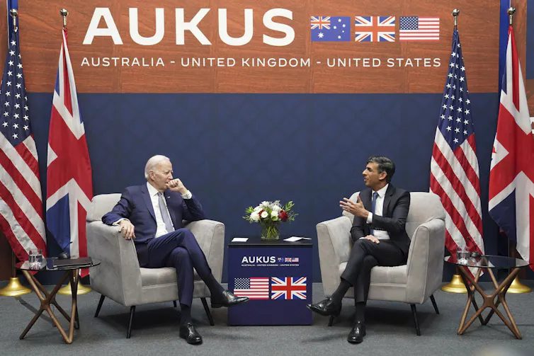 Two men sit in easy chairs talking with an AUKUS sign behind them and flags next to them.