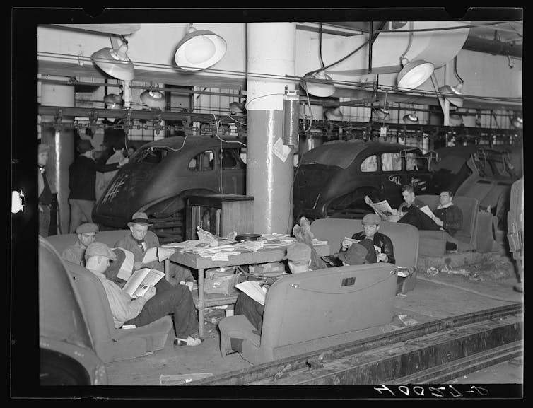 In a black-and-white photo, several striking autoworkers read newspapers, sitting on car seats placed on the ground like sofas. They ignore the unfinished chassis behind them.
