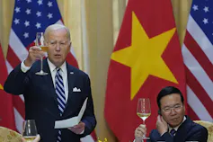 An older man stands with a raised champagne glass as a younger man sits next to him also raising a glass.