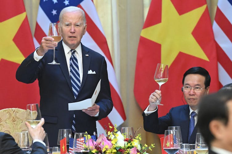 A gray haired man holds paper in his left hand as he raises a half-filled champagne glass in his right hand. To his left, a seated man wearing glasses raises his half-filled champagne glass.