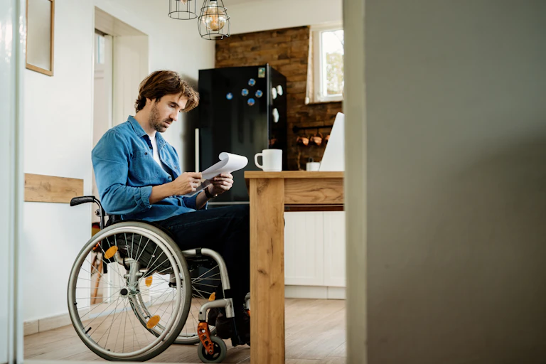 A man in a wheelchair looks at some paperwork at a kitchen table