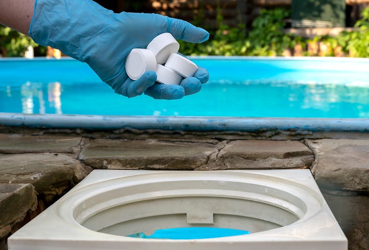 A hand in a blue glove tossing several white tablets into a reservoir next to a swimming pool