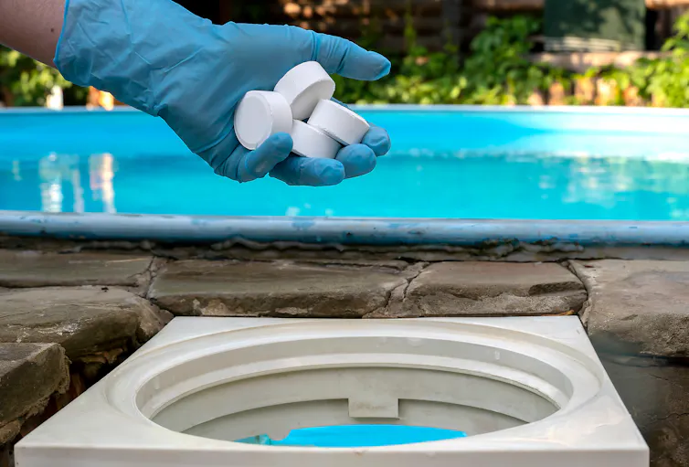 A hand in a blue glove tossing several white tablets into a reservoir next to a swimming pool
