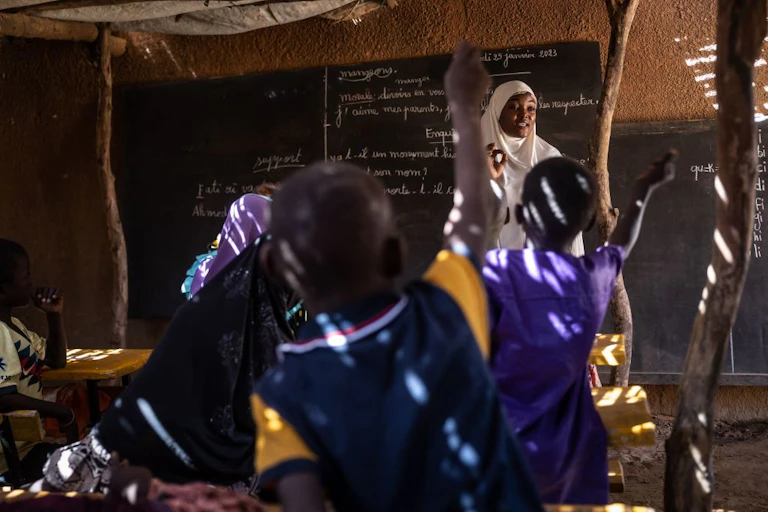 A woman wearing a white hijab stands in front of a chalkboard while her pupils raise their hands