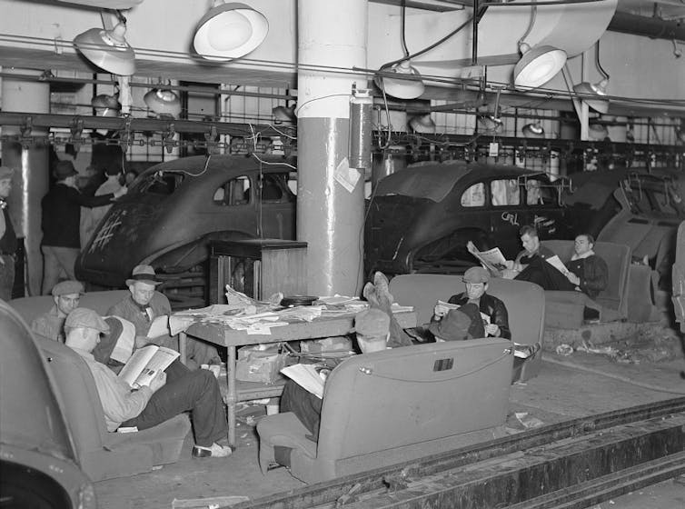 In a black-and-white photo, several striking autoworkers read newspapers, sitting on car seats placed on the ground like sofas. They ignore the unfinished chassis behind them.