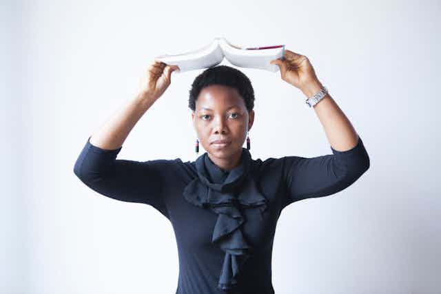 A young African woman wearing a black blouse with frills holds a paperback book open above her head.