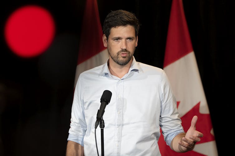 A man in a white shirt speaks into a microphone. A Canadian flag is behind him.