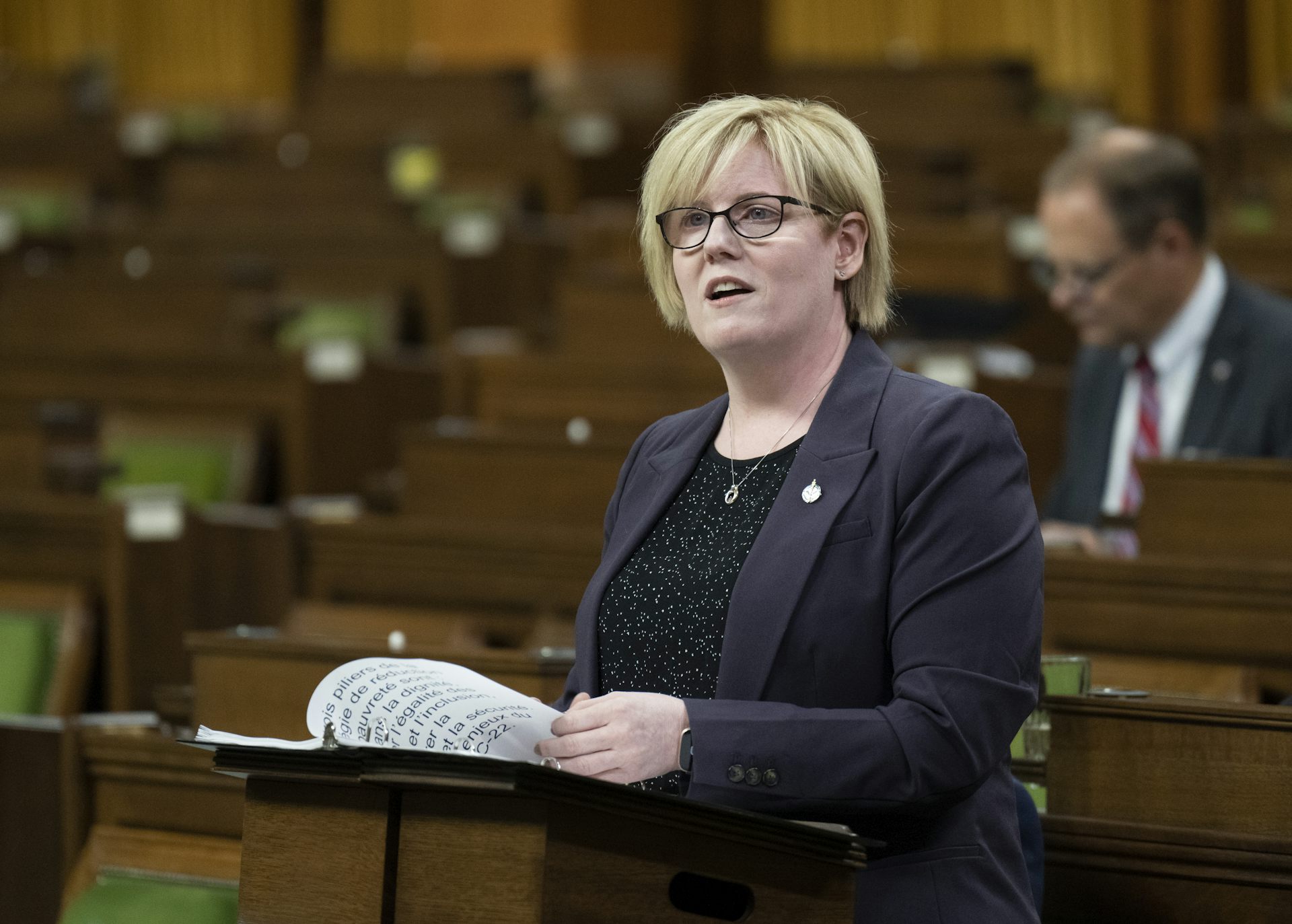A middle-aged woman with short, blonde hair and glasses speaks from behind a podium