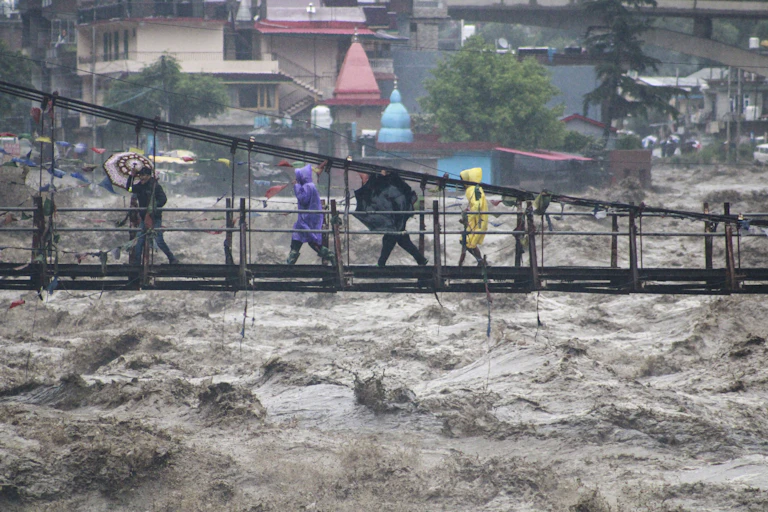 People walk across a foot bridge over raging water with umbrellas and raincoast sheltering against a strong rainfall.