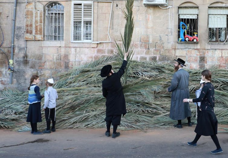 A man in black clothes holds up a tall palm branch he has selected from a large pile of them on the street.