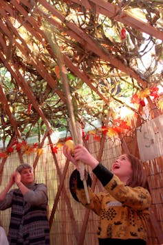 A young girl looks up toward the roof of a hut, smiling, as she holds a branch and a yellow fruit.