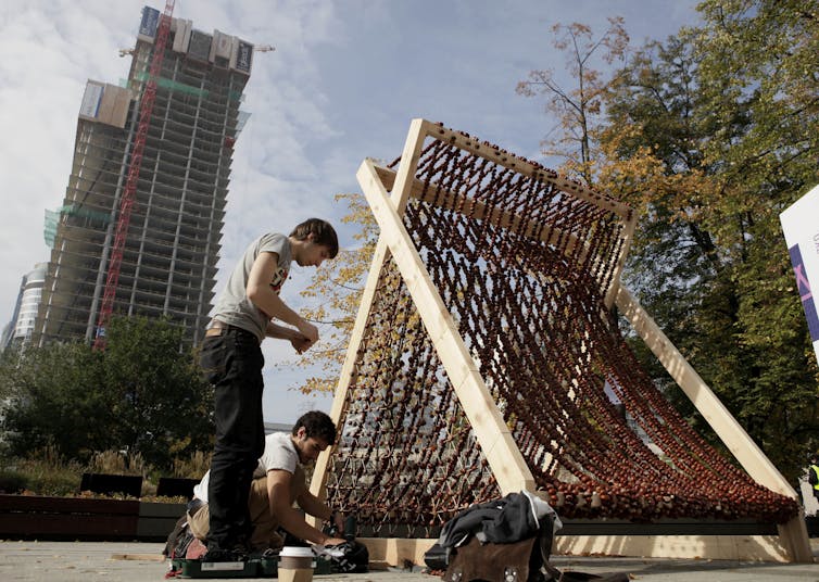 Two men in casual clothing construct a wooden fort in a city plaza.