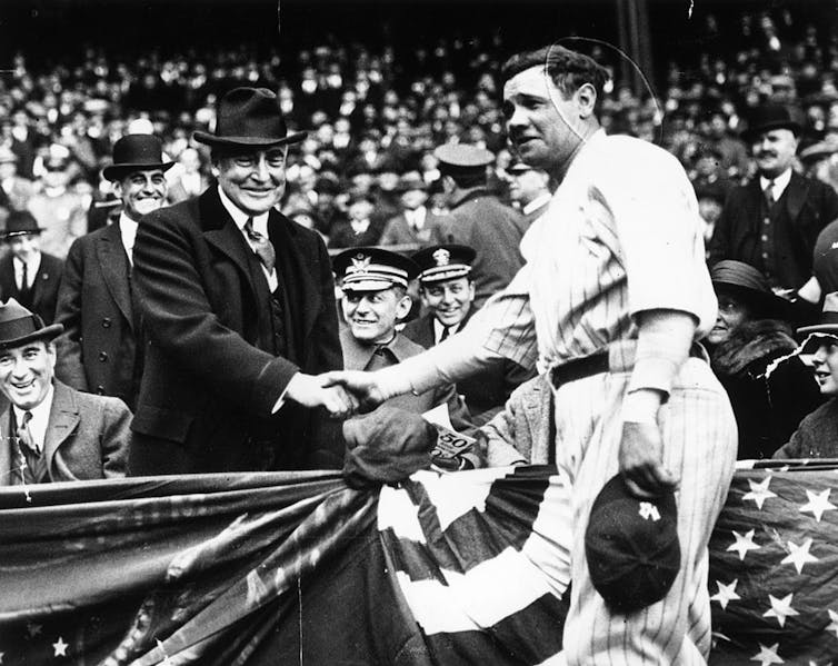 A man in a white baseball uniform with a baseball glove on one hand shakes hands with a man in a dark coat and fedora.