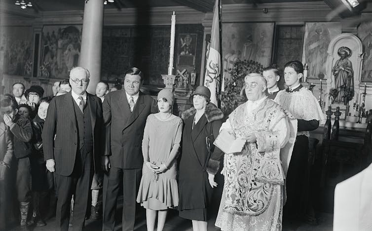 Half a dozen people pose in a row, wearing formal clothing. One of them is a priest wearing a stole.