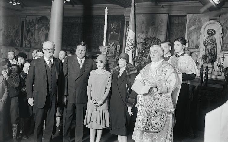Half a dozen people pose in a row in formal wear. One of them is a priest who wears a stole.