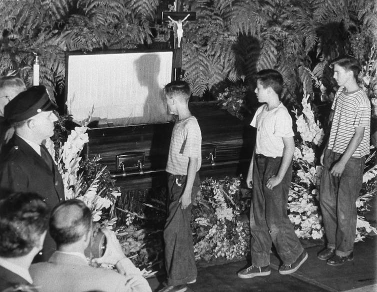 Three preteen boys in t-shirts and pants walk solemnly past an open casket surrounded by plants.